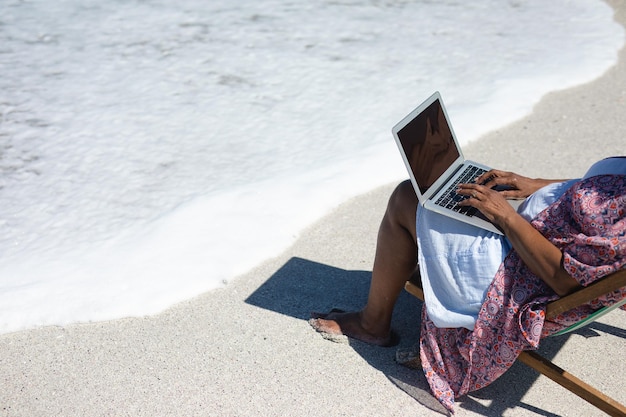 Vista de alto ângulo de uma mulher afro-americana sênior em uma praia ao sol, sentada em uma cadeira de terra, usando um computador portátil