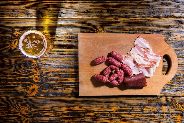 Vista de alto ângulo de uma tábua de cortar com salsichas, bacon e carne na mesa de madeira perto de um copo cheio de cerveja