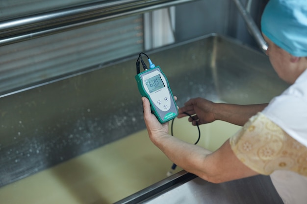 Vista de alto ângulo de uma trabalhadora madura segurando o medidor de temperatura enquanto examina a mistura de queijo em uma fábrica de alimentos industrial, copie o espaço