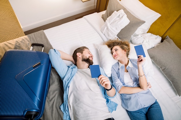 Vista de alto ângulo do jovem casal alegre de turistas, deitada na cama no quarto de hotel, segurando seus passaportes. Mala azul na cama