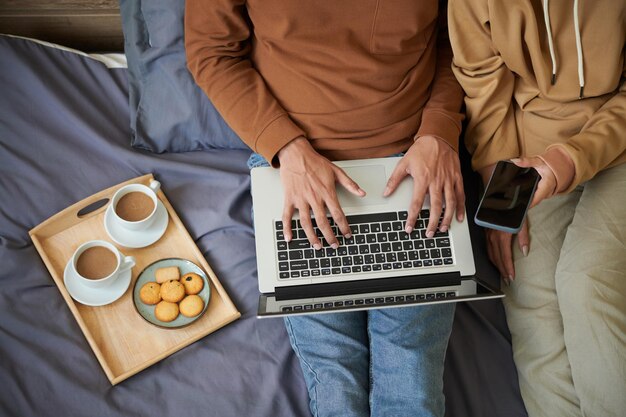 Vista de alto ângulo do jovem casal sentado na cama e usando seus gadgets para fazer compras on-line durante o café da manhã