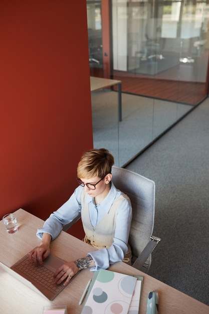 Vista de alto ângulo em uma moderna mulher de negócios tatuada usando o laptop na mesa enquanto trabalha no escritório com a parede vermelha, copie o espaço