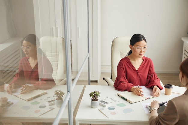 Vista de alto ângulo para uma empresária asiática moderna falando com o cliente enquanto trabalha na mesa em um cubículo de escritório branco, copie o espaço