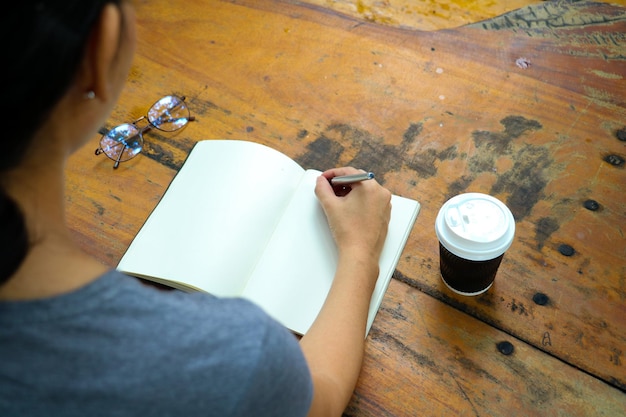 Vista de ângulo alto de mulher escrevendo em livro por xícara de café e óculos na mesa