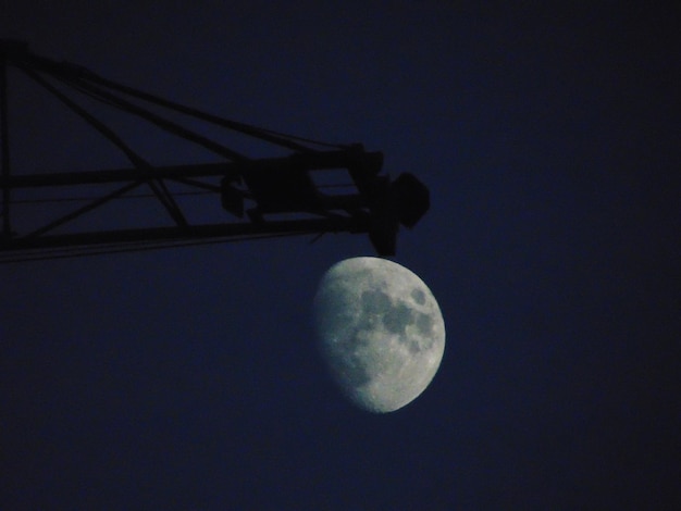 Foto vista de ângulo baixo de guindaste em silhueta por lua gibosa contra o céu à noite