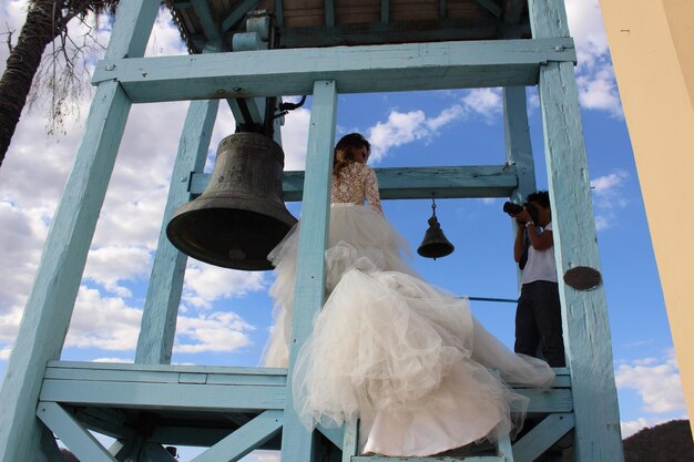 Vista de ângulo baixo de homem fotografando noiva de pé ao lado do sino