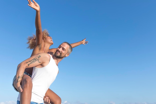 Vista de ângulo baixo de homem piggybacking namorada alegre contra o céu azul