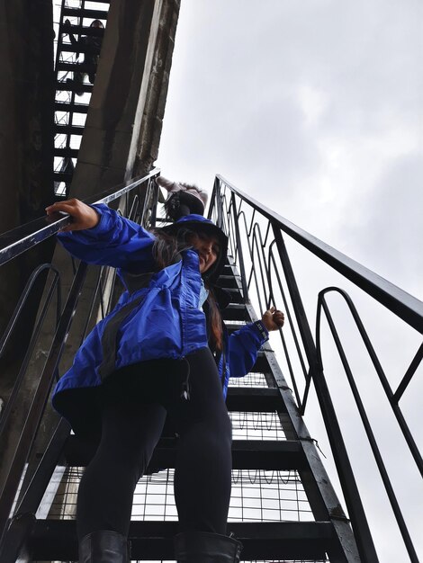 Foto vista de ângulo baixo de mulher vestindo uma jaqueta azul de pé na escada contra o céu