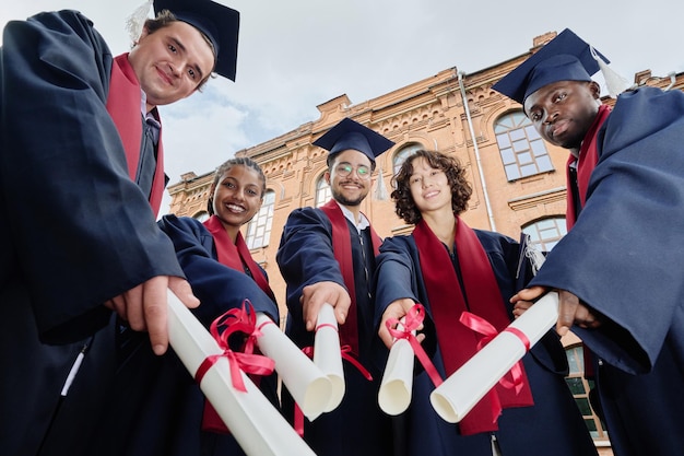 Vista de ângulo baixo de um grupo de estudantes graduados com diplomas sorrindo para a câmera enquanto estão ao ar livre