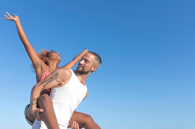 Vista de ângulo baixo de um homem carregando uma namorada alegre contra um céu azul claro