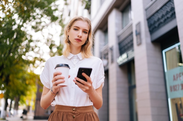 Vista de ângulo baixo de uma jovem muito confiante em roupas casuais, bebendo café para viagem e usando smartphone andando na rua urbana