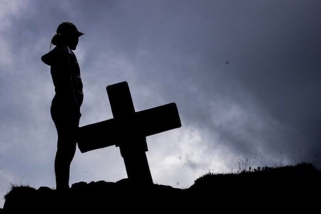 Foto vista de ângulo baixo de uma mulher em silhueta de pé junto a uma cruz religiosa em uma colina contra o céu