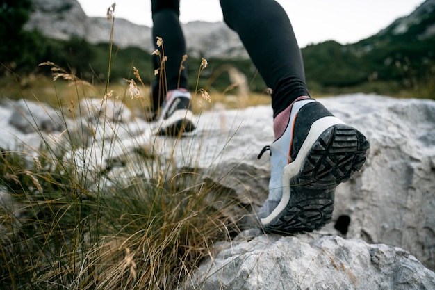 Vista de baixo ângulo através da grama de montanha crescendo das rochas de sapatos de caminhada femininos pisando ou subindo uma trilha de montanha rochosa