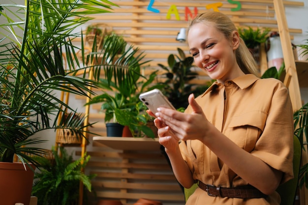 Vista de baixo ângulo da alegre florista feminina digitando mensagem para o cliente e conversando com o cliente usando o celular olhando para a tela do smartphone