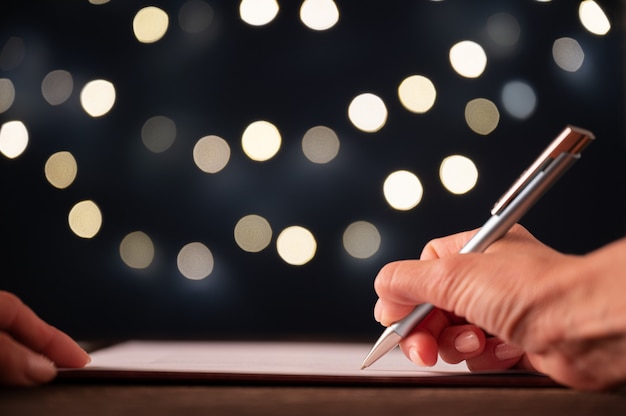 Vista de baixo ângulo da mão feminina, assinando um documento, contrato ou resolução com as luzes borradas do feriado no fundo.