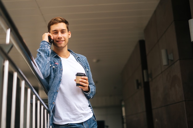 Vista de baixo ângulo de um jovem sorridente falando no celular, em pé no terraço da varanda com uma xícara de café de café encostada na grade do prédio de escritórios, fundo desfocado, olhando para a câmera.