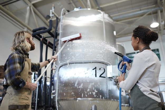 Vista de baixo ângulo para dois trabalhadores lavando equipamentos em uma cervejaria industrial moderna, copie o espaço