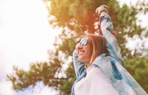 Vista de baixo de uma jovem mulher bonita e feliz com óculos escuros e camisa xadrez azul, levantando os braços sobre um céu e árvores. Liberdade e aproveite o conceito.
