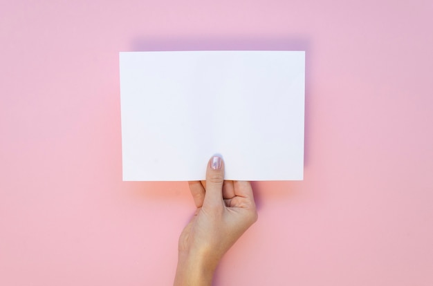 Vista de cima, a mão feminina segura a maquete da folha de papel em branco sobre um fundo rosa pastel.