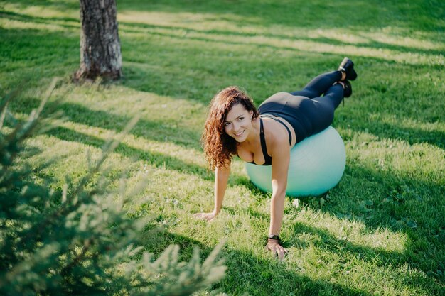 Vista de cima da mulher morena alegre faz exercício de pilates com bola de fitness tem poses de treinamento esportivo ao ar livre na grama verde no parque aproveita o dia ensolarado Mulher realiza ioga lá fora