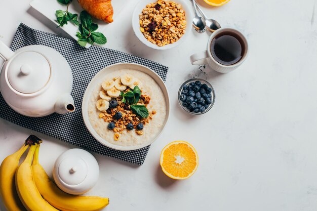 Foto vista de cima de uma mesa branca com farinha de comida com frutas e nozes frutas bagas chá hortelã laranjas bananas pratos de chá com espaço livre