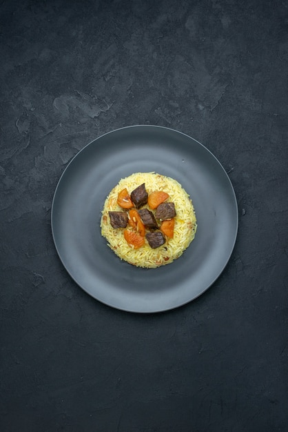 Vista de cima, delicioso pilaf cozido de arroz com damascos secos e fatias de carne dentro do prato em superfície escura