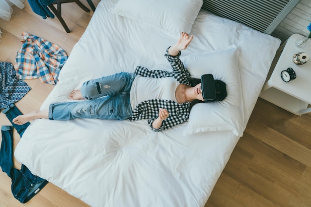 vista de cima despreocupada garota asiática em roupas casuais está deitada na cama se divertindo usando fone de ouvido vr no quarto com roupas espalhadas no chão em casa.