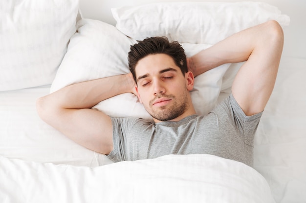 Vista de cima do homem musculoso morena de camiseta casual, dormindo sozinho em casa na cama com lençóis brancos, colocando as mãos sob a cabeça