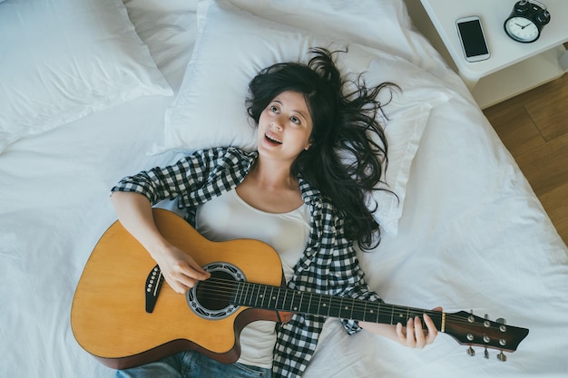vista de cima feliz músico feminino asiático está cantando e tocando um violão enquanto estava deitado na cama de lazer no quarto em casa.
