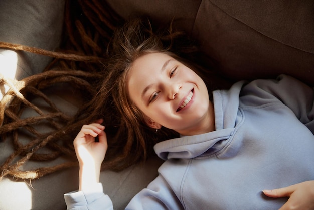 Vista de cima retrato de jovem sorridente com dreadlocks longos em capuz azul deitado no sofá