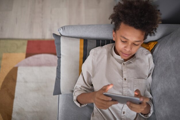 Vista de cima, retrato médio de um menino adolescente relaxando no sofá em casa jogando videogame no smartphone, copie o espaço