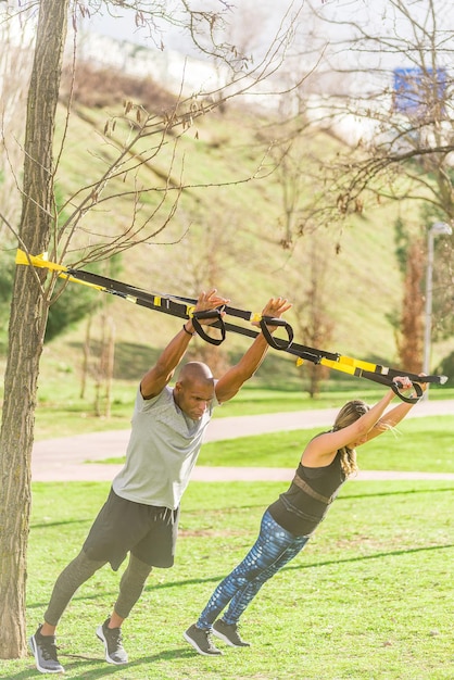 Foto vista de corpo inteiro de casal fitness fazendo exercícios de braço com alças trx fitness no parque. pessoas multiétnicas fazendo exercícios ao ar livre.