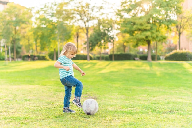 Vista de corpo inteiro de um menino brincando com uma bola na grama no parque em um dia ensolarado.