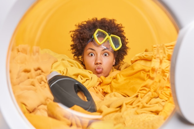 Foto vista de dentro da máquina de lavar cheia de roupa. mulher engraçada e encaracolada com máscara de mergulho na testa