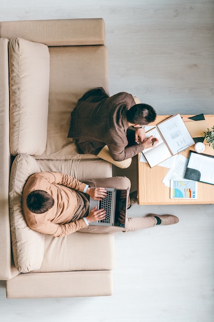 Vista de dois jovens empresários sentados em um sofá macio, um deles fazendo anotações enquanto seu colega navegava na rede