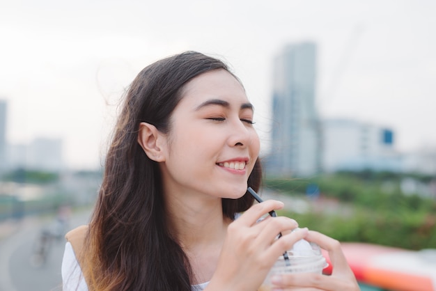 Vista de perfil de mulher jovem e atraente com uma xícara de café na mão, sentado ao ar livre e apreciando a vista da cidade moderna.