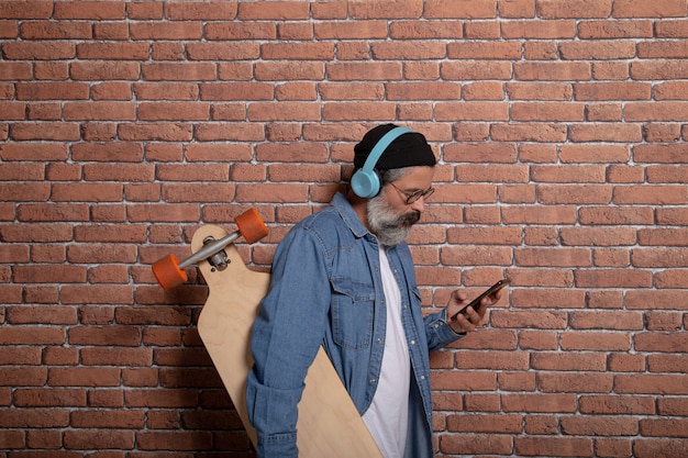 Vista de perfil de um homem moderno com barba, chapéu, fones de ouvido e longboard olhando para o telefone na parede de tijolos