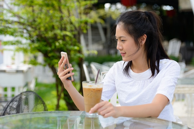 Vista de perfil de uma bela jovem asiática usando o telefone em uma cafeteria ao ar livre