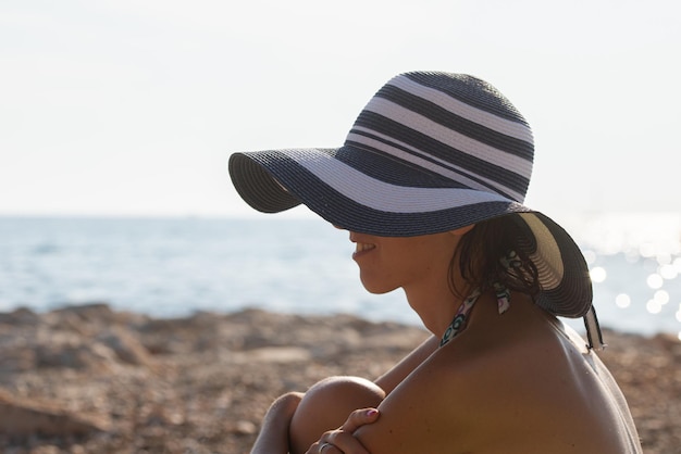 Vista de perfil de uma jovem com chapéu de sol sentado em uma praia rochosa em um dia de verão.