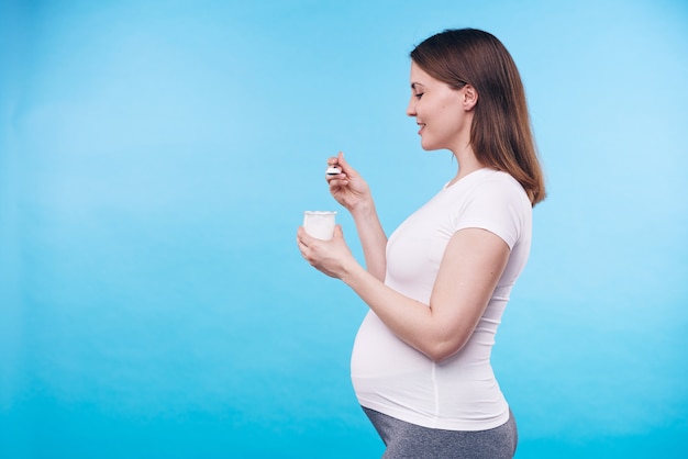 Vista de perfil de uma jovem grávida saudável em uma camisa branca comendo iogurte em um pequeno copo de plástico em pé na parede azul