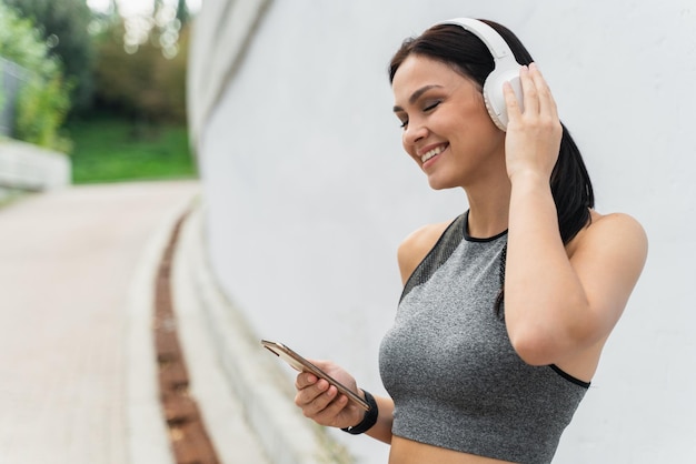 Vista de retrato da cintura para cima da jovem sorridente atlética em roupas esportivas cinza e fones de ouvido grandes, ficando no estádio e curtindo a música