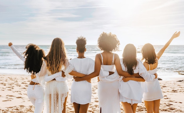 Vista de trás de senhoras multirraciais em vestidos brancos posando na praia e olhando para o oceano tendo donzela