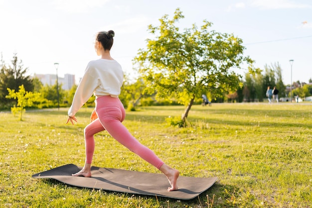 Vista de trás de um treino feminino irreconhecível antes de uma sessão de treinamento de fitness no parque em pé em um tapete de ioga Mulher jovem saudável aquecendo-se ao ar livre esticando as pernas Conceito de estilo de vida saudável