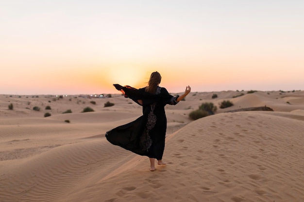 Vista de trás de uma linda mulher misteriosa em um tradicional vestido longo preto árabe fica no deserto ao pôr do sol