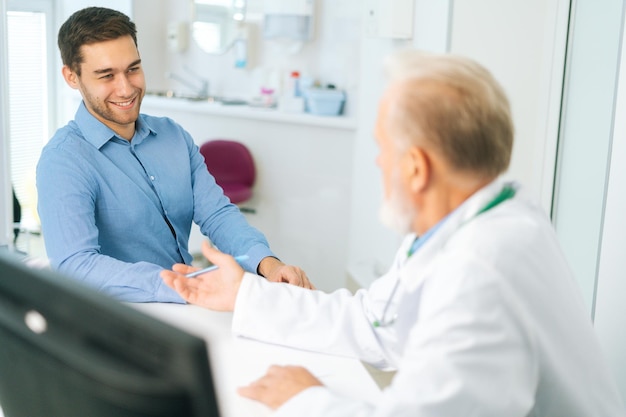 Foto vista de trás para o paciente jovem alegre ouvindo médico sênior dando conselhos de saúde no hospital médico sênior entrevistando jovem doente e escrevendo prescrição