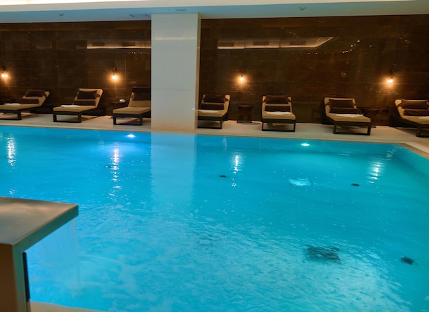 Vista de um belo e espaçoso interior com piscina térmica coberta com cascata, iluminações subaquáticas e espreguiçadeiras em uma luxuosa área de estar de um resort spa de bem-estar
