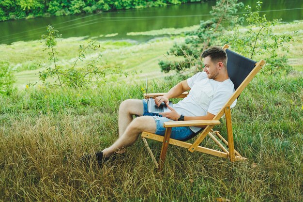 Vista de um cara bonito deitado na cadeira na natureza e trabalhando com laptop descansando sozinho olhando pensativamente para cima Um homem em uma viagem no campo o conceito de estilo de vida das pessoas