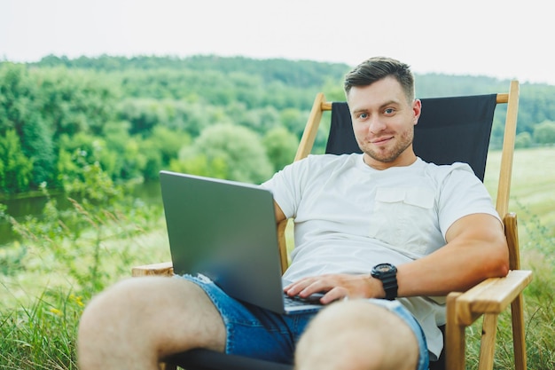 Vista de um cara bonito deitado na cadeira na natureza e trabalhando com laptop descansando sozinho olhando pensativamente para cima Um homem em uma viagem no campo o conceito de estilo de vida das pessoas