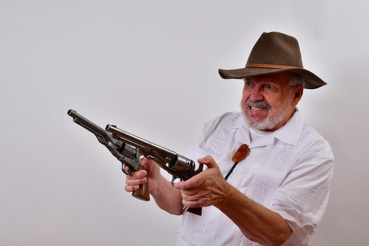 Foto vista de um homem idoso com um chapéu segurando duas armas no ar com um rosto louco em branco