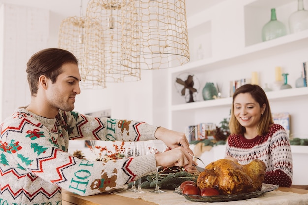 Vista de um jovem e uma mulher sentados em uma mesa decorada de Natal, prontos para comer um peru assado